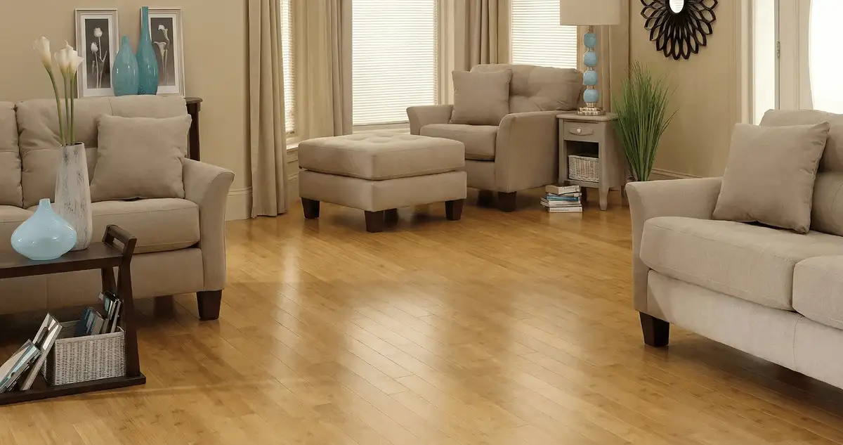 A living room with professionally installed bamboo flooring, beige sofas, a matching ottoman, a side table with a blue lamp, vases, framed pictures, and large windows with blinds and curtains in Birmingham, AL